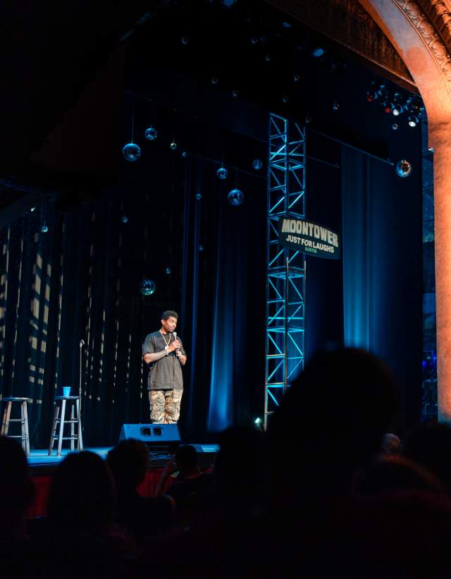 Deon Cole performing stand-up comedy on stage at Paramount Theater during Moontower Just for Laughs Festival in Austin with dramatic lighting and vintage theater backdrop.