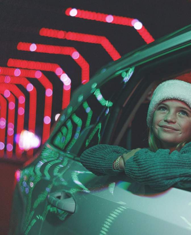 A young girl wearing a Santa hat looks out a car window at a festive drive-through Christmas light display during a holiday vacation in Branson, Missouri