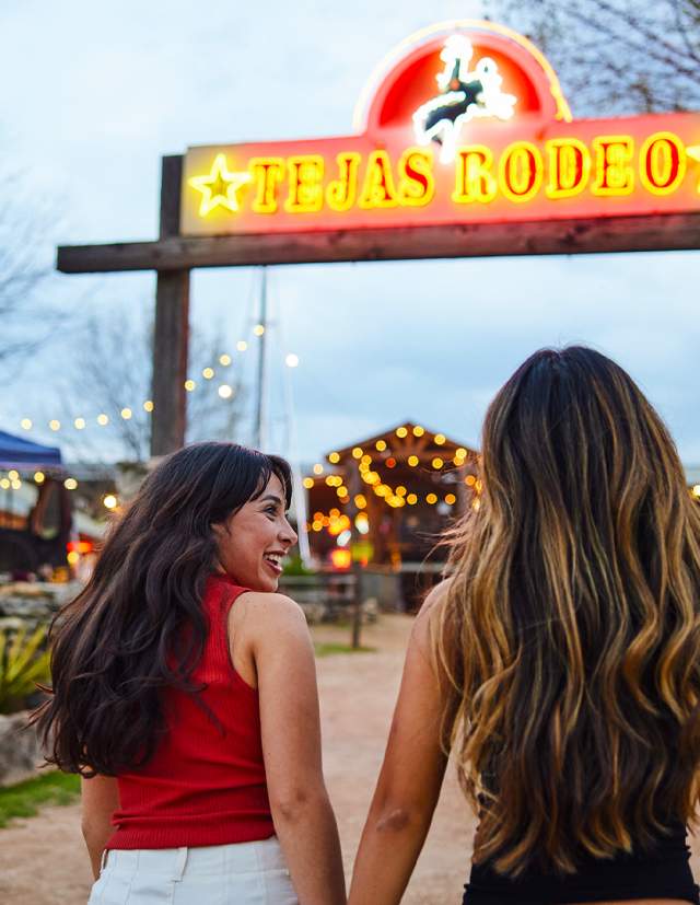 Two girls walking under neon sign that reads "Tejas Rodeo."