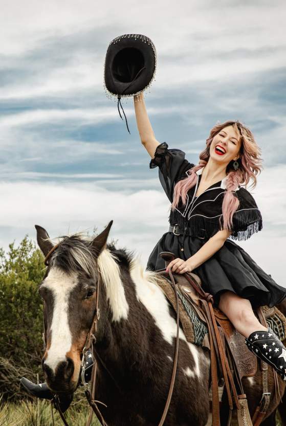 a woman in a black fringe dress and star-spangled cowboy boots rides a paint horse through the wide-open grasslands near Amarillo, raising her black hat high with a joyful smile