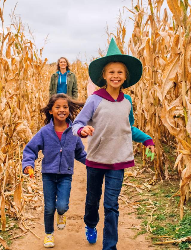 Two girls, once with witch hat, run through corn maze with mom in the background