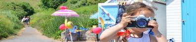 A young girl in a white and blue dress holds a camera, posing playfully. Behind her are colourful beach huts and umbrellas, set in lush greenery.