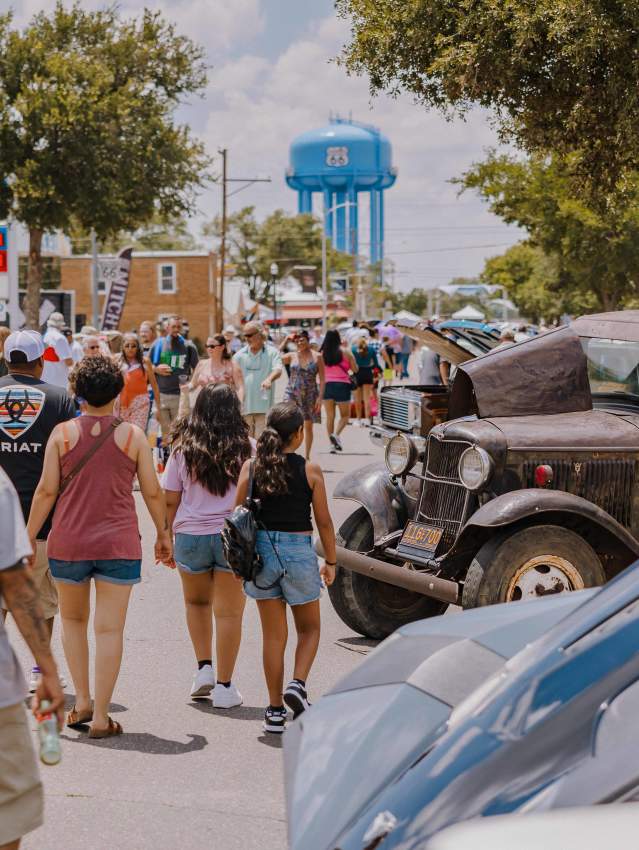 Crowds stroll past vintage cars on display under the summer sun in Amarillo’s Historic Route 66 District, with the bright blue Route 66 water tower rising in the background. Festival flags wave above taco stands and classic car hoods, capturing the lively spirit of the Texas Route 66 Festival finale.