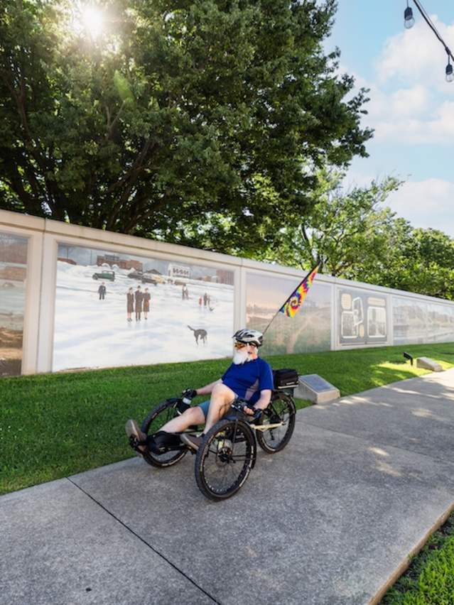A man pedals a recumbent bike past Paducah's Wall to Wall Floodwall murals. A woman is in the background on a bicycle. The sun shines through the trees.