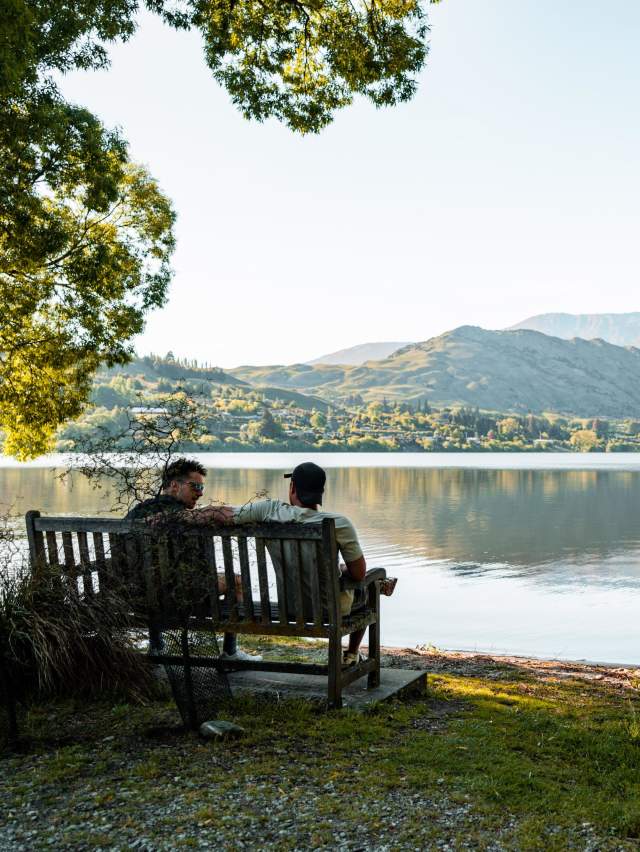 Two people sitting on a bench looking our over a lake with mountains in the background