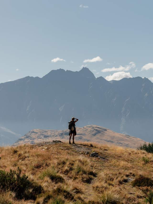 Person at the top of the Queenstown Hill summit with mountains behind them