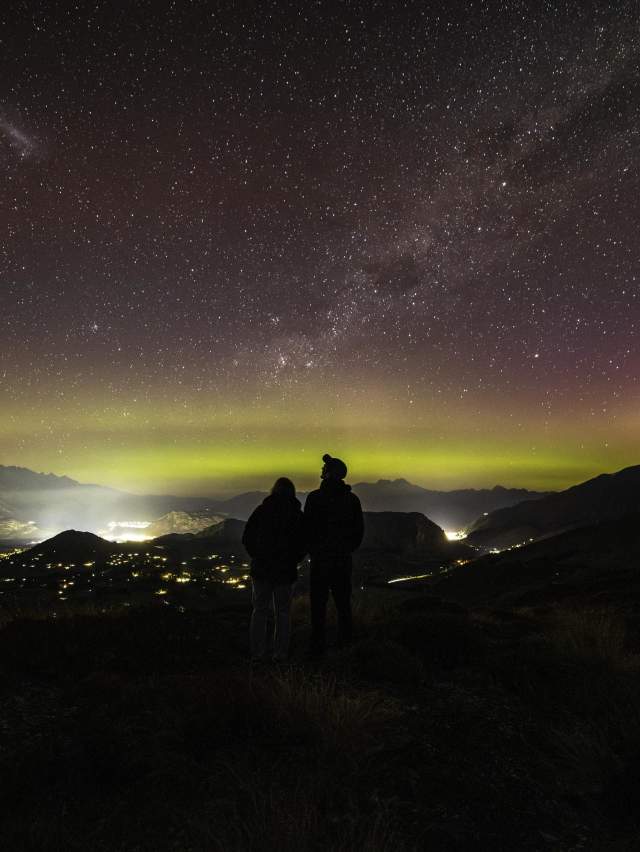 People standing on a mountain top with a starry night sky and aroura before them