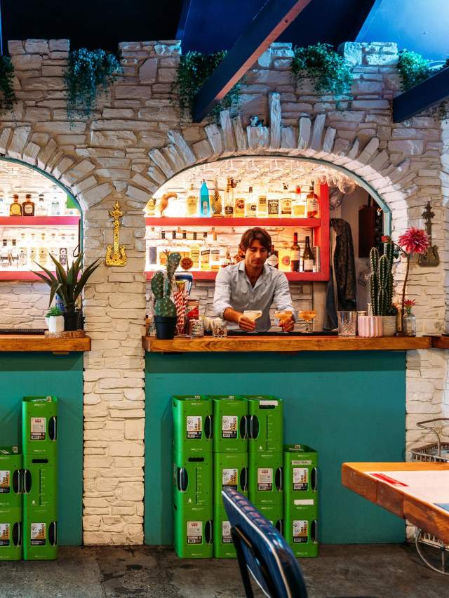 A vibrant Mexican-inspired bar with stone archways, colourful decor, and a bartender crafting drinks behind a wooden counter.