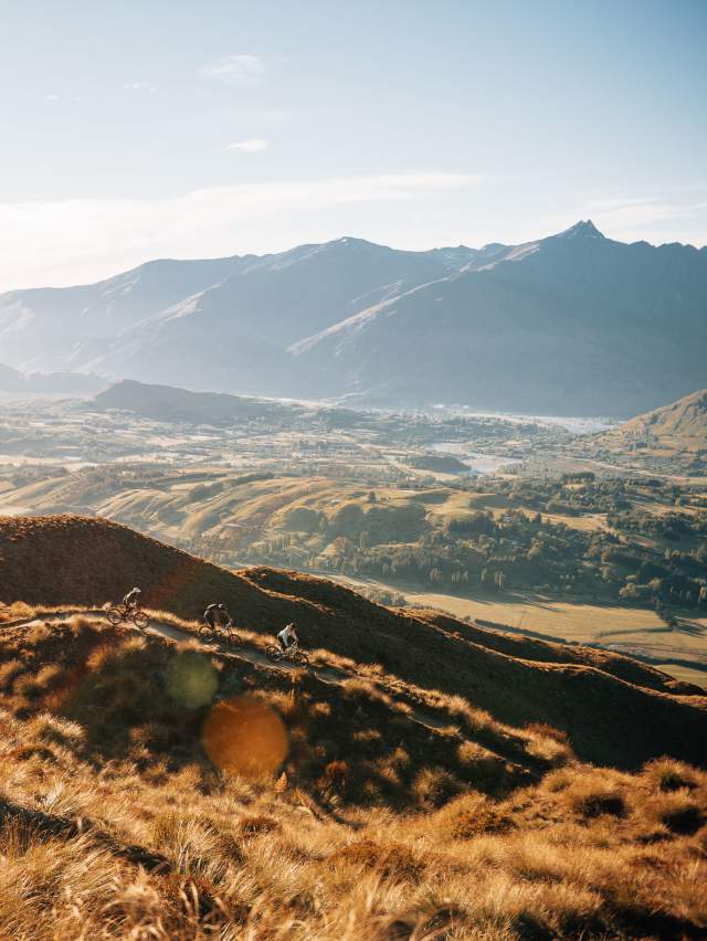 Far shot of three bikers descending a golden tussock-covered mountain with valley and mountain in the background