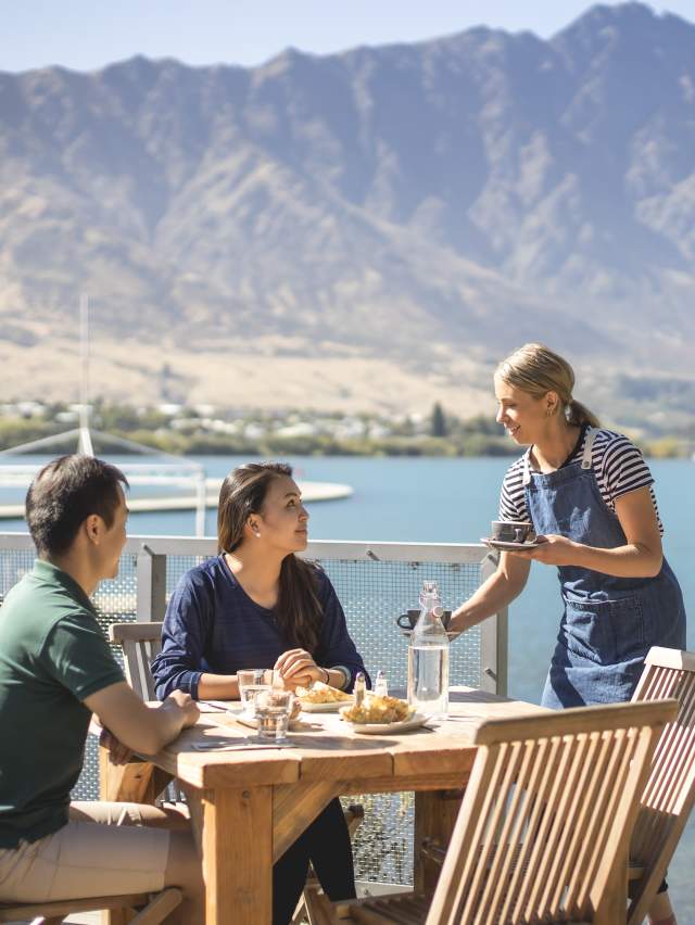 Couple being served by a waitress outside at the Boat Shed café, with the Remarkables Mountains in the background