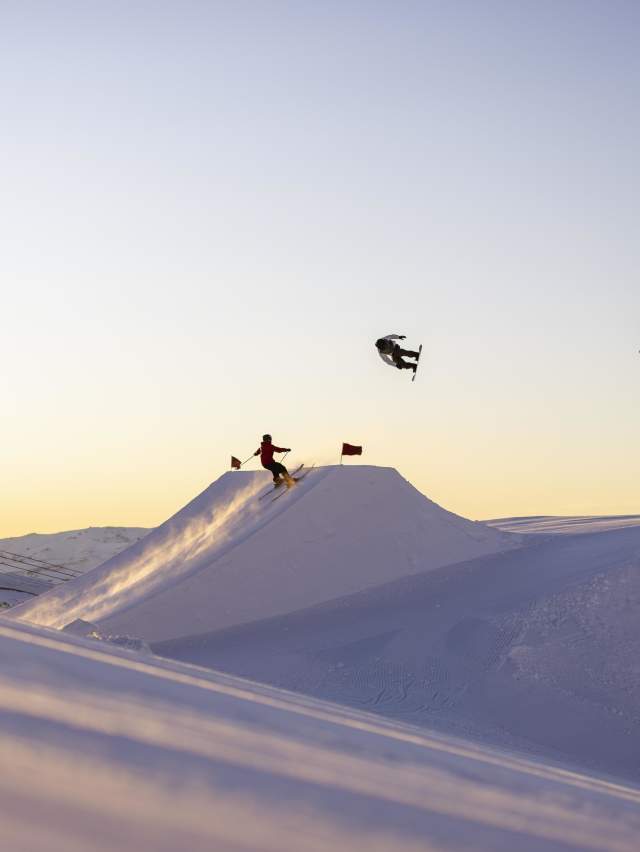 Snowboarders and a skier perform aerial tricks off a jump at Cardrona Alpine Resort at sunrise.