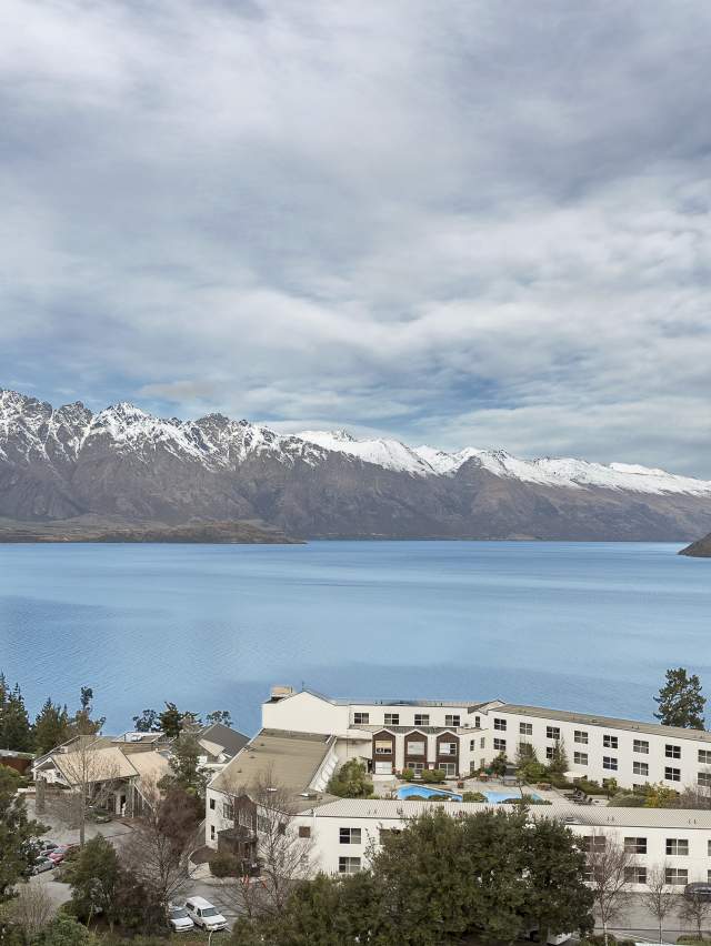A stunning view of Lake Wakatipu with the Remarkables mountain range in the background, featuring a lakeside hotel and surrounding greenery in the foreground.