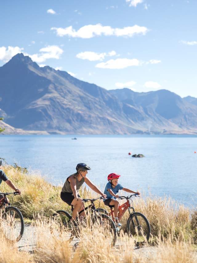 A Family biking around the Queenstown Gardens, with Lake Whakatipu and Cecil Peak in the background