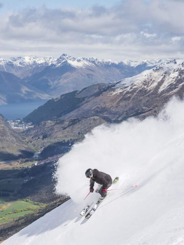 Person snowboarding down a mountain with views of green valley and mountains in the background