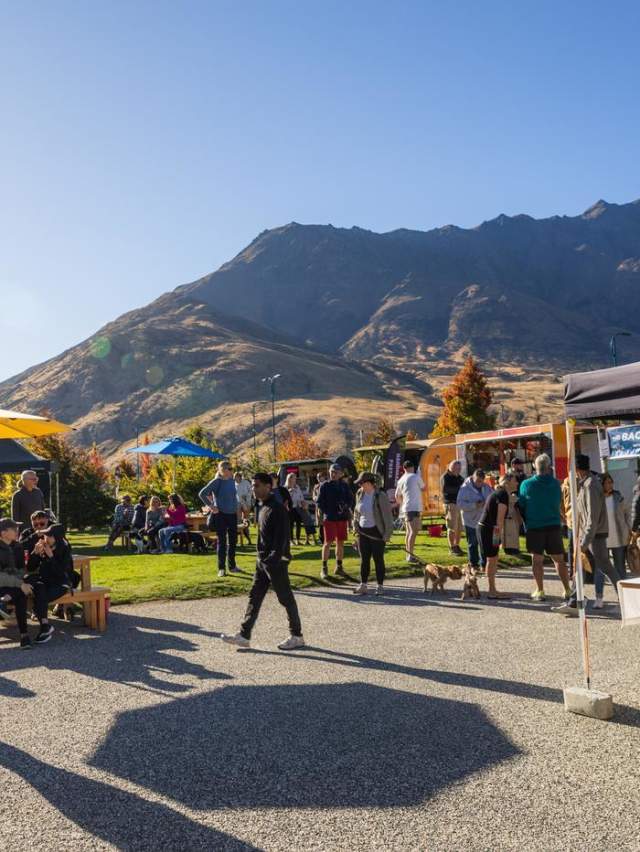 People browsing stalls and relaxing under colourful umbrellas at the Remarkables Market, with mountains in the background on a sunny day.