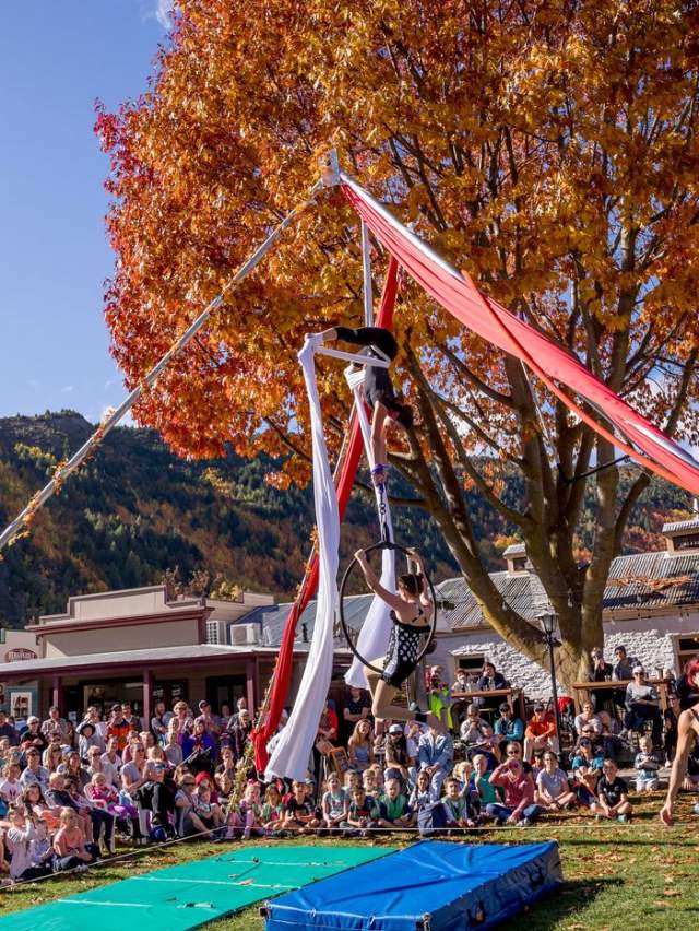 Crowd gathered around an aerial performance sounded by red and gold trees at the Arrowtown Autumn Festival