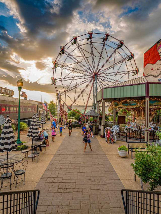 A colorful photo of the Park at West End in Downtown Fort Smith, featuring a ferris wheel, nickel & dime shop, Native American mural, adults, and children.