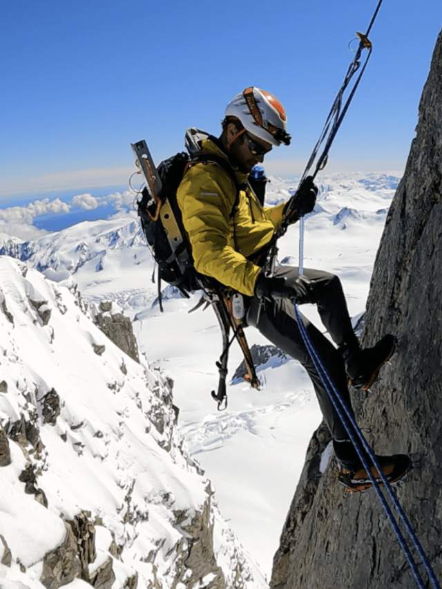 Mountaineer Hamish Fleming rappelling down a rock face with snow-covered mountains in the background