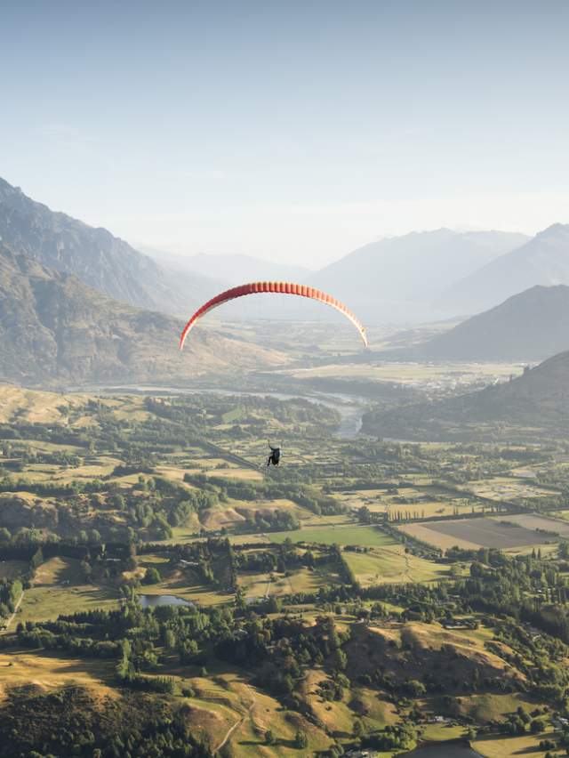 Person paragliding over a green valley with the Remarkables mountain range in the background