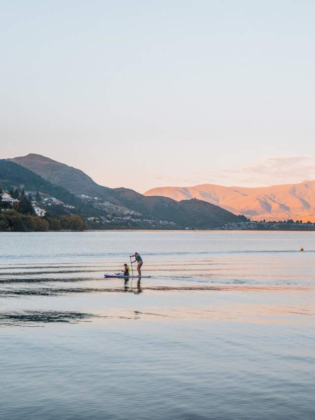 Paddle boarder in middle of still lake at dusk with mountains in the background