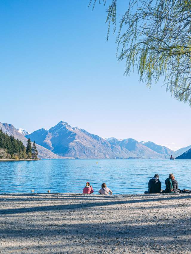 People sitting on Queenstown beach in spring with lake views and mountains in the background