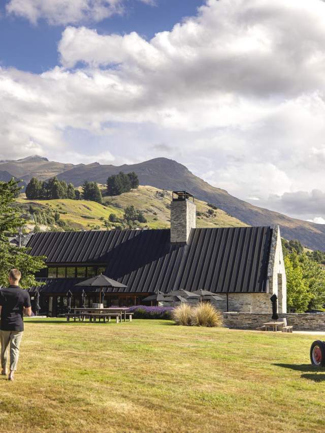 Two people walking along a lawn towards a restaurant with views of mountains in the background