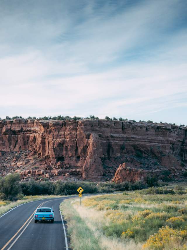 A car drives on a winding road past a large red rock formation under a clear blue sky.