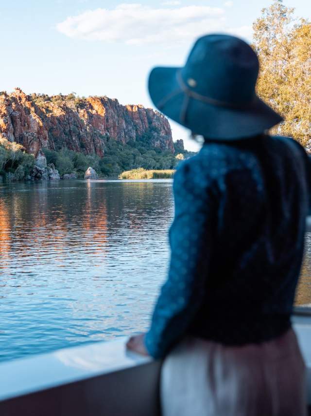A woman enjoying the scenery on an Ord River cruise - the woman is standing on the vessel looking across the water at a cliff faced bathed in sunlight.