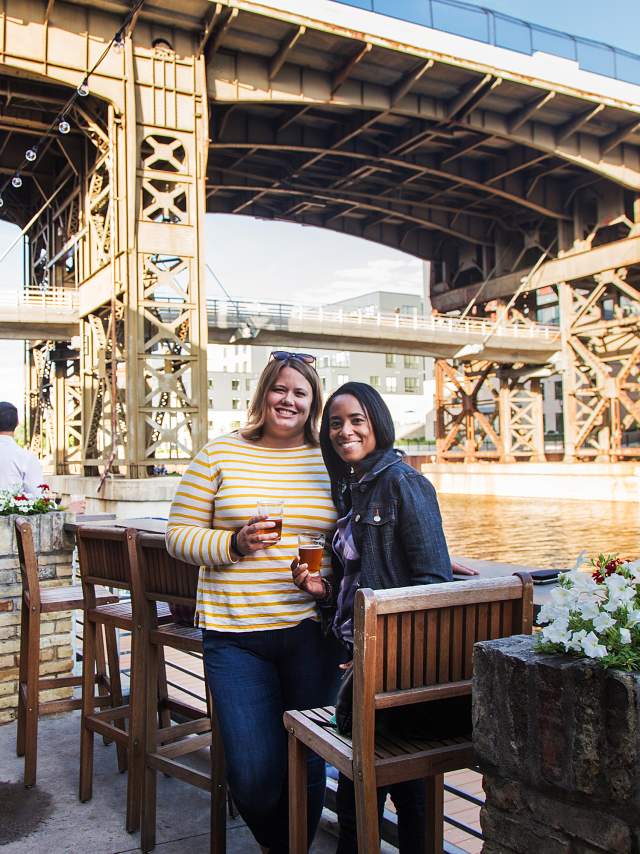 People enjoying drinks at the outdoor riverwalk, Lakefront Brewery under a large steel bridge on a sunny day.