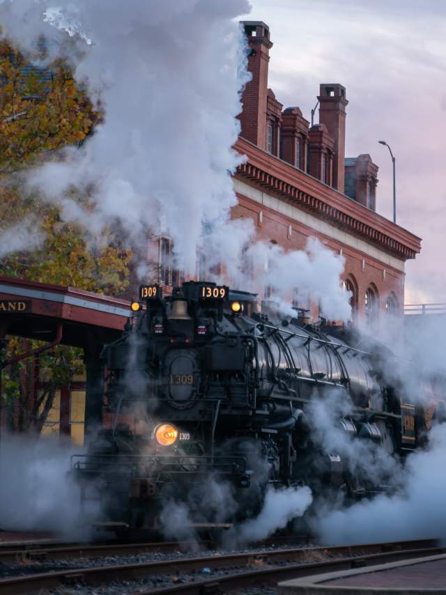 A large black steam train engine pulls into the Cumberland Station as it releases a billowy cloud of steam.