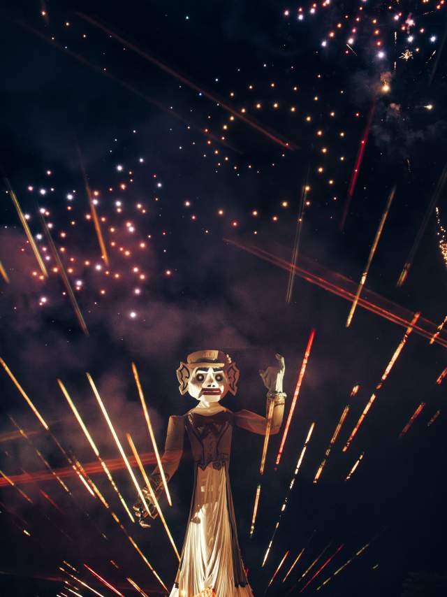 A large effigy of Zozobra is lit up against a night sky filled with colorful fireworks. Bright streaks of light and sparks are scattered around, enhancing the festive atmosphere.