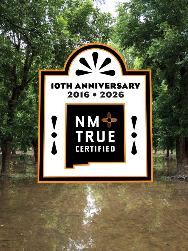 A forested area with partially submerged trees in floodwater is shown. There is a large logo displayed in the center, celebrating the 10th anniversary of "NM True Certified" from 2016 to 2026.