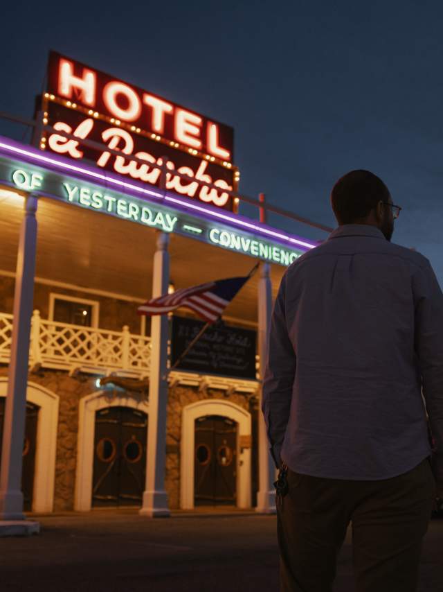 Two people standing in front of a hotel named "Hotel El Rancho," which is lit up at dusk with a sign that reads "Charm of Yesterday, Convenience of Today." An American flag hangs prominently on the building.