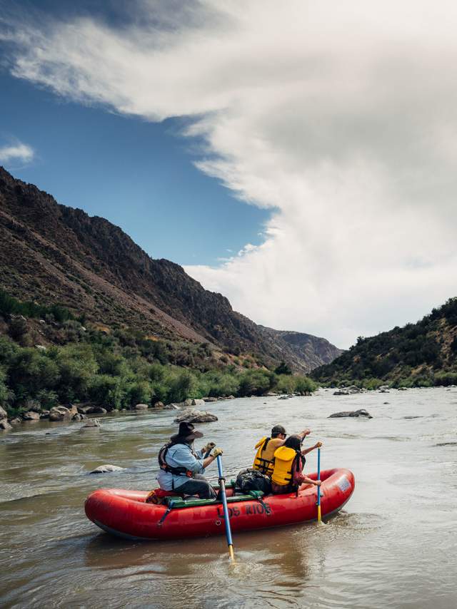 two people sit in a red raft as they paddle down a flowing river between two sprawling hills on either side
