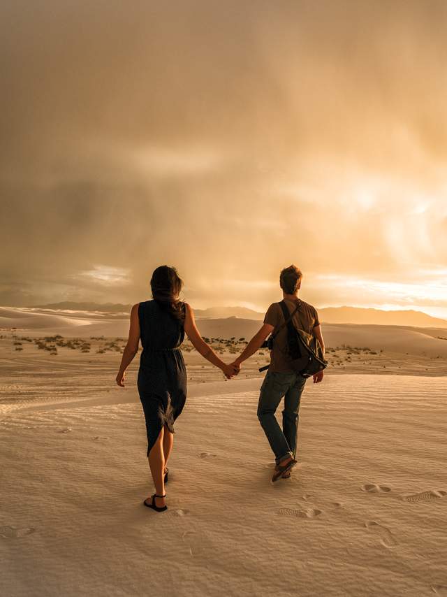 two people hold hands while walking across white sand towards a glowing sunset amongst fluffy white clouds