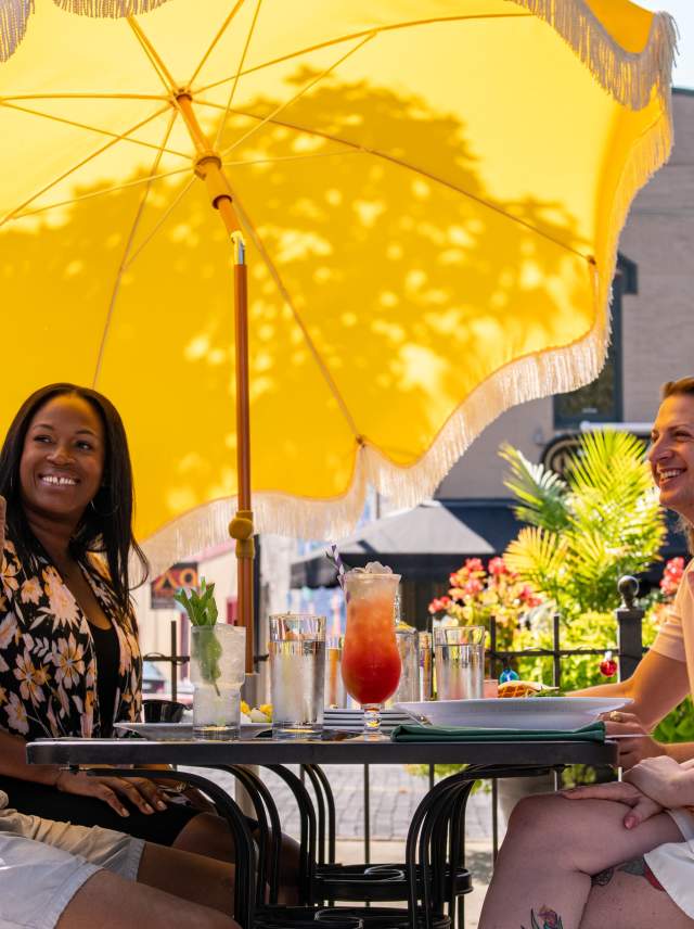 A group of people sitting at an outdoor patio table, under a large yellow umbrella