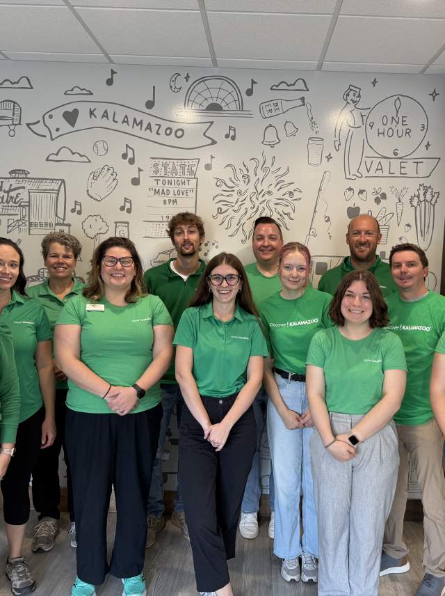 The team of Discover Kalamazoo all wear green shirts, standing in front of the lobby mural.