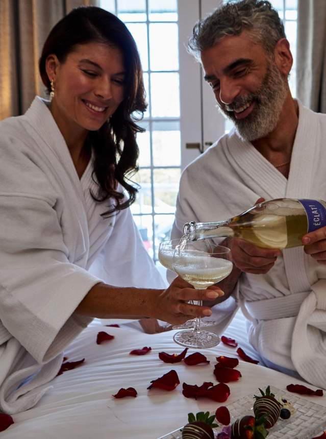 Couple pouring sparkling wine in a hotel bedroom. Rose petals and chocolate strawberries on bed.