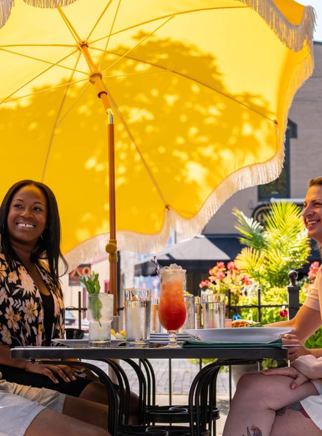 A group of people sitting at an outdoor patio table, under a large yellow umbrella