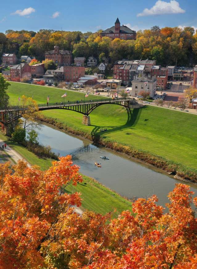 Drone image of the Galena River, a walking bridge, and downtown Galena. Autumn leaves of orange, gold, and green surround the town.