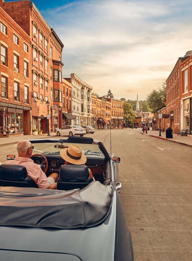 Two people riding in a blue convertible car, down a Main Street of brick buildings.