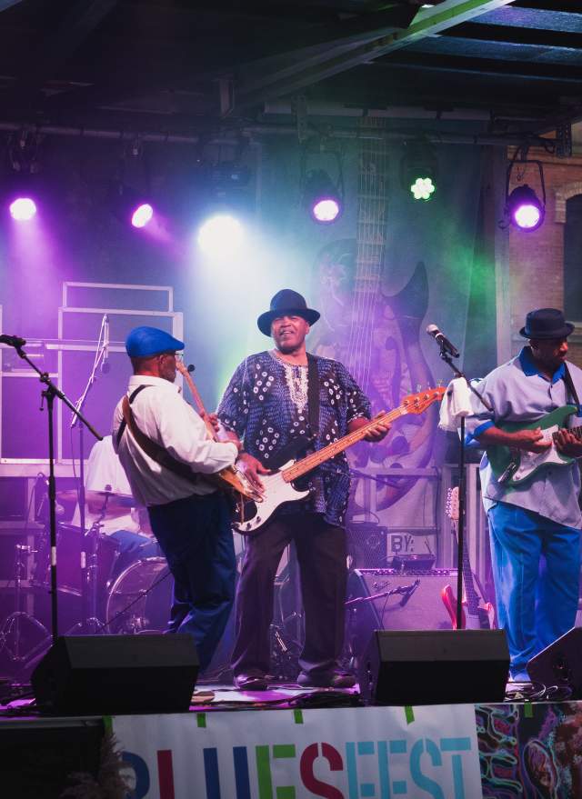 A blues band performing on an outdoor stage at Michigan BluesFest, with guitars, drums, and a cheering crowd.