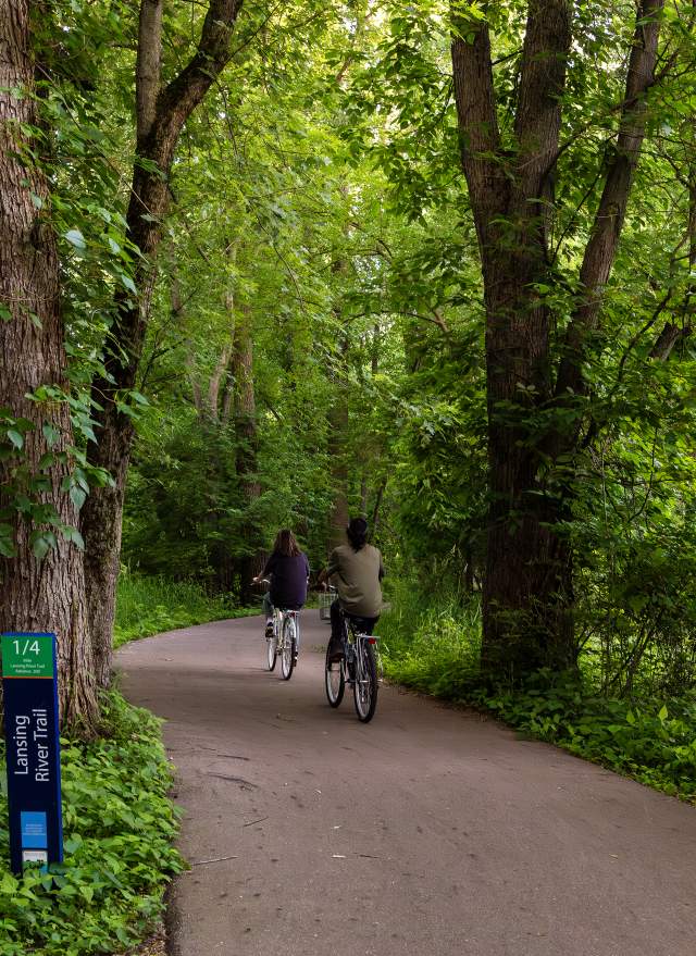 Two bikers riding on the Lansing River Trail surrounded by lush green plants