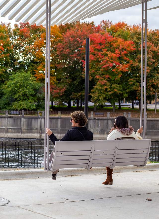 Two people sitting on a bench swing overlooking the Grand River while another person takes their photo