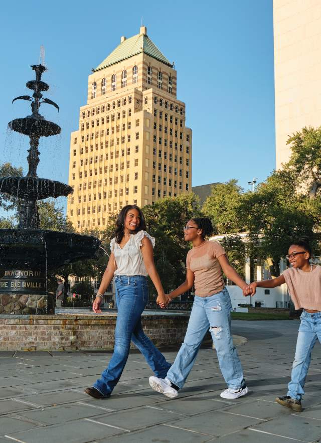 Two Black girls and one Black boy walk in a park past a fountain with buildings in the background