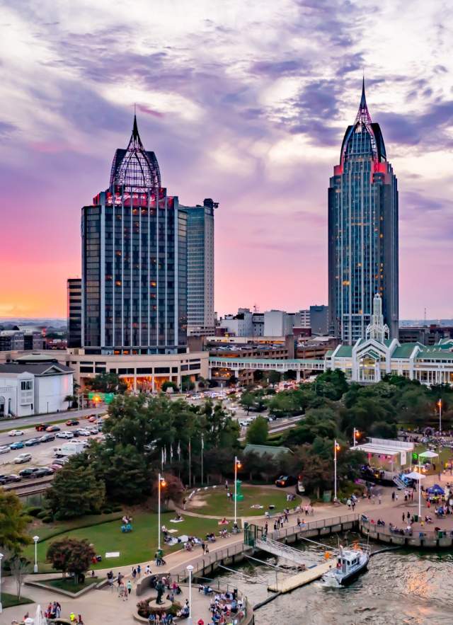 A vibrant sunset over Downtown Mobile skyline, featuring tall buildings, a bustling waterfront, and a lively crowd below.