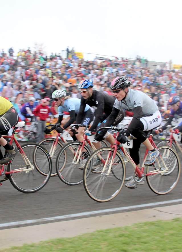 Cyclists in helmets race on the track during the Little 500 bike event with a large crowd watching from the stands in the background.