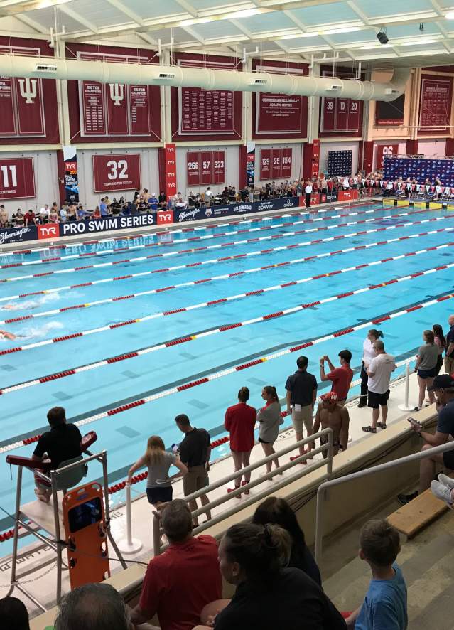 Bloomington indoor swimming pool with lane dividers, red and white banners, and people standing at the edge.