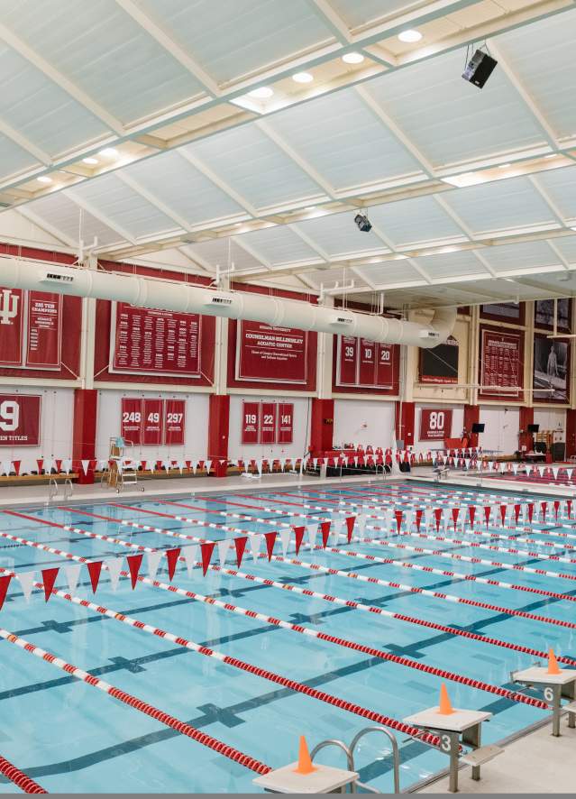 Bloomington indoor Olympic-sized swimming pool with lane dividers, red and white banners, and three people standing at the edge wearing lifeguard uniforms.