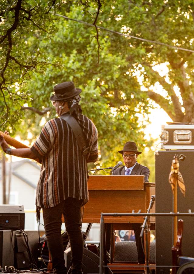 Booker T. Jones (on the piano) and his band members. The image is taken from the back of the stage and there is music equipment and speakers throughout the stage.